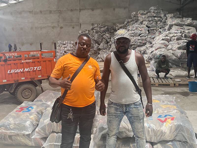 two men in a warehouse with bales of used clothing on the background